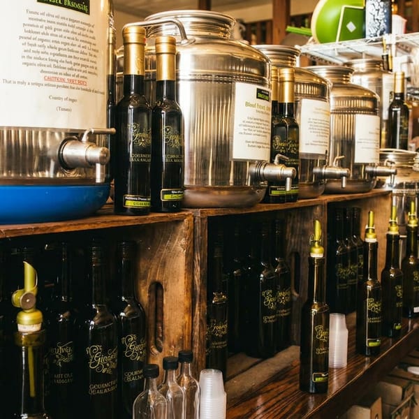 Stainless steel olive oil dispensers and bottled oils on wooden shelves in a specialty store.