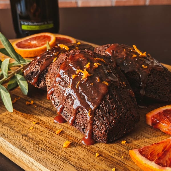 Chocolate loaves with orange glaze and zest, blood orange slices, and olive branch on a wooden board.