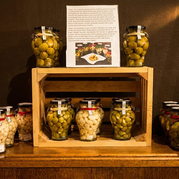 Jars of olives and garlic cloves displayed on a wooden shelf with a descriptive sign.