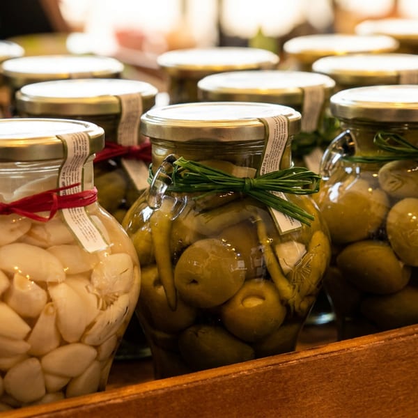 Jars of pickled garlic cloves and green olives with decorative ribbons on a wooden shelf.